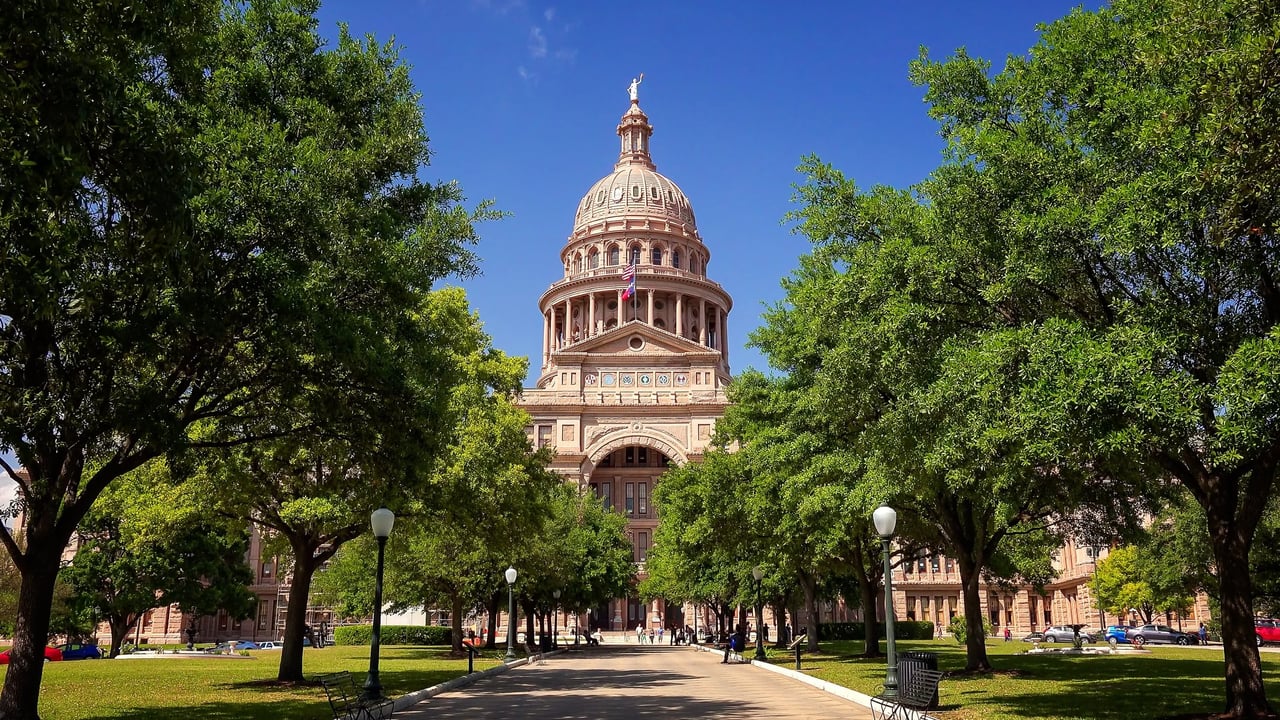 The Texas State Capitol rises beyond a shaded, tree-lined walkway.