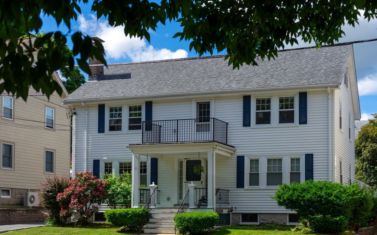White two-story house with blue shutters, a covered entry porch, and a small upper balcony.