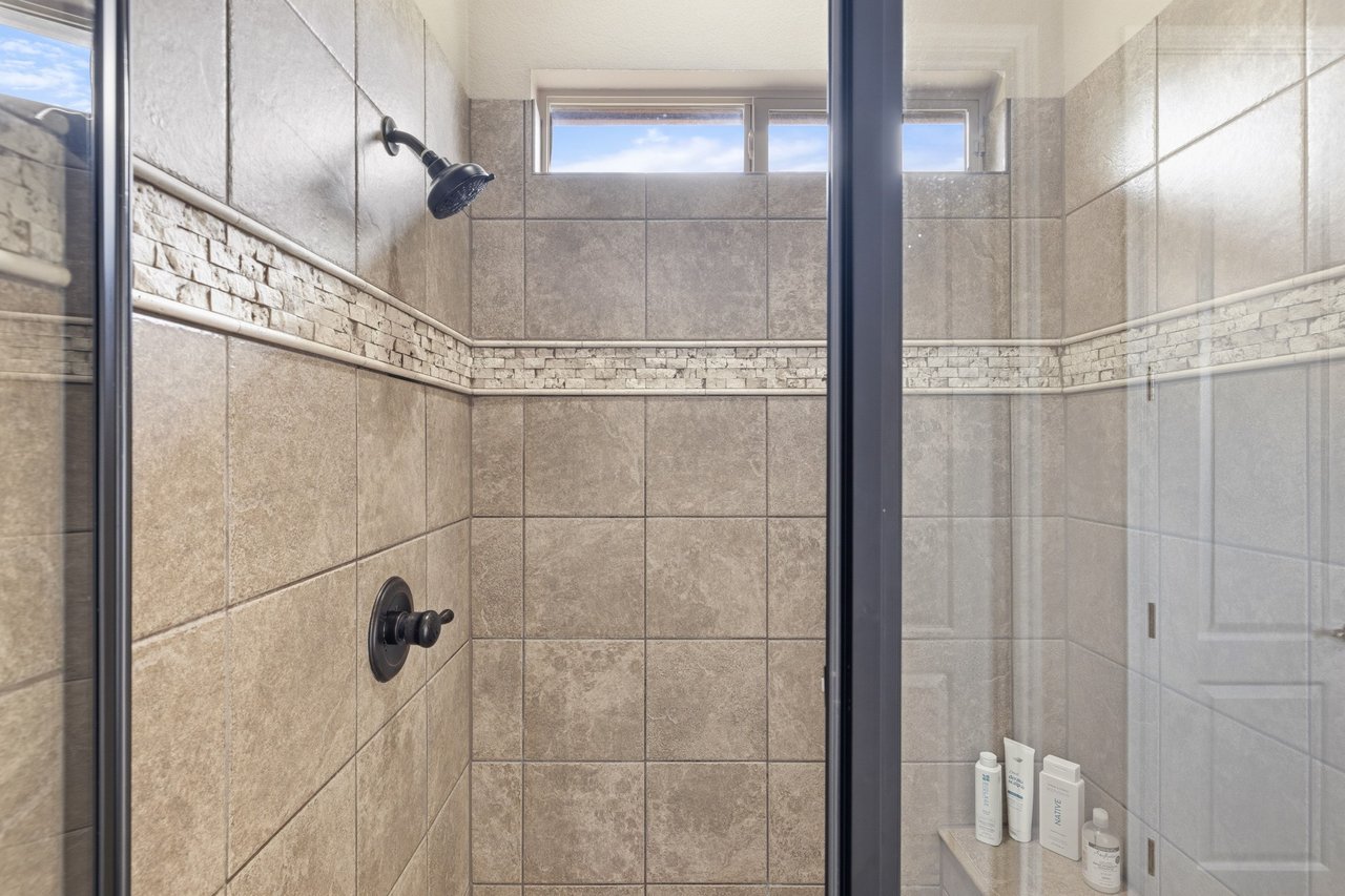 Walk-in shower with stone-look tile, a mosaic accent band, dark fixtures, and high windows.