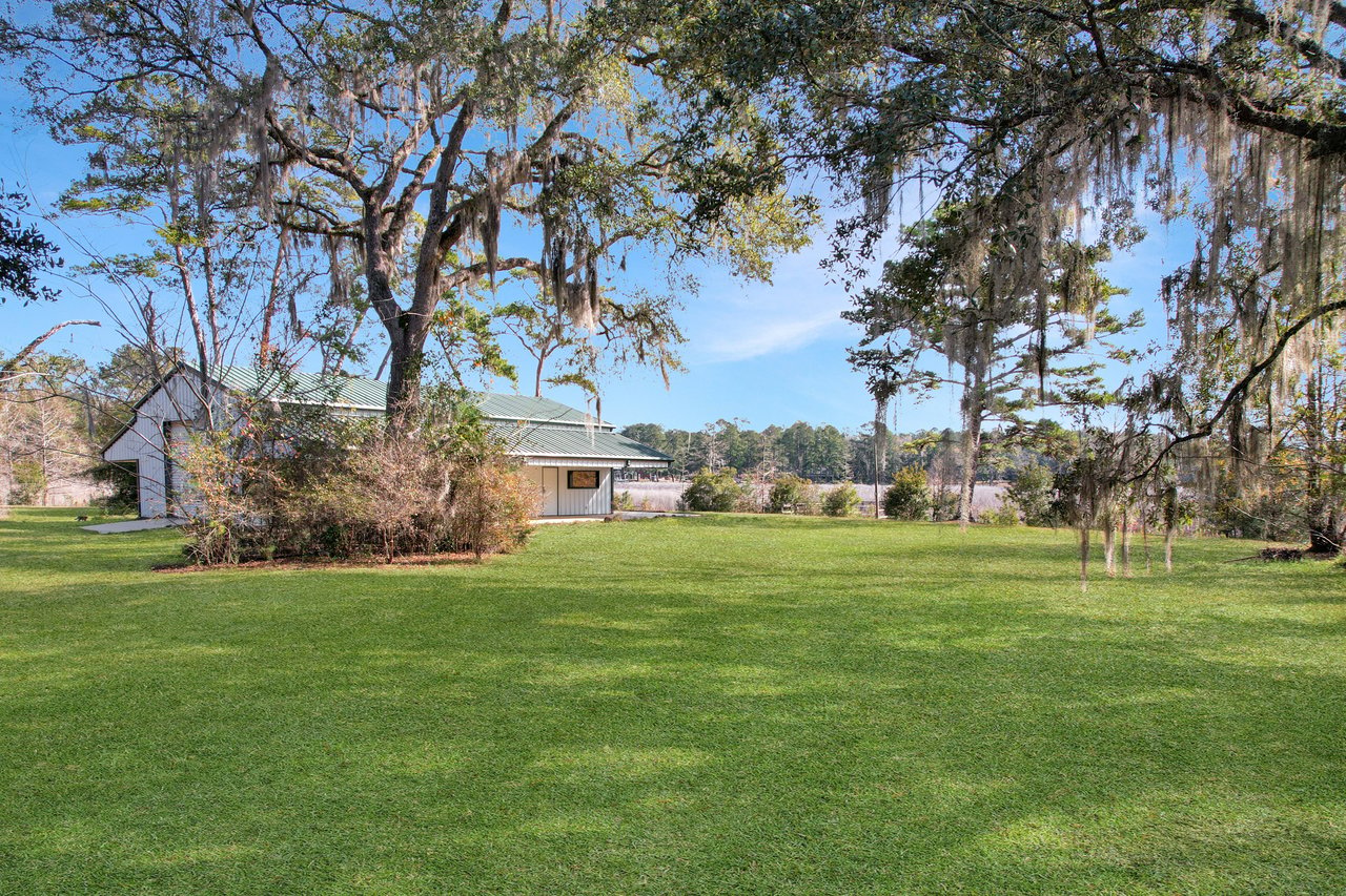 Sprawling green lawn with large oak trees draped in Spanish moss. A white house with a green roof is partially visible, set against a clear blue sky.