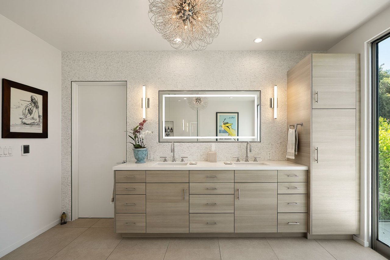 Modern double-sink bathroom with pale wood cabinetry, a large mirror, and a sculptural chandelier.