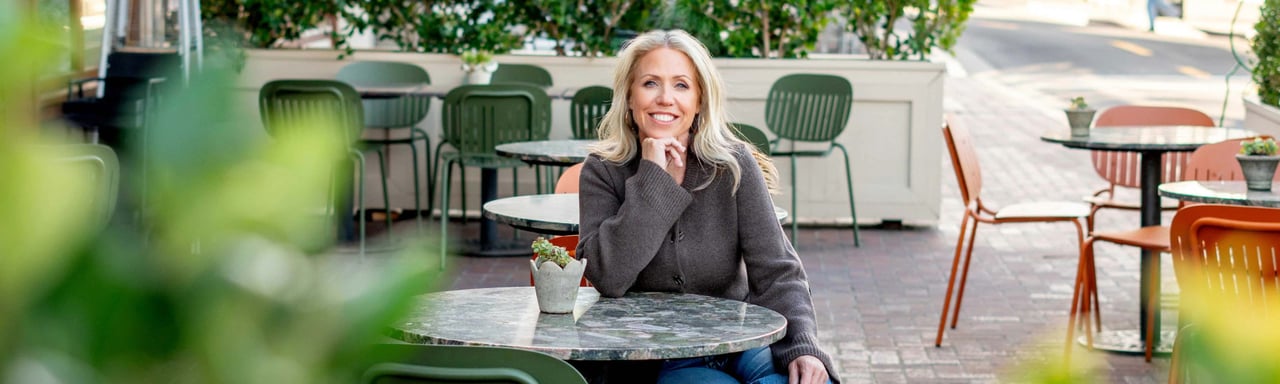 Maile Mock smiling at a patio table, framed by leafy plants and café seating.