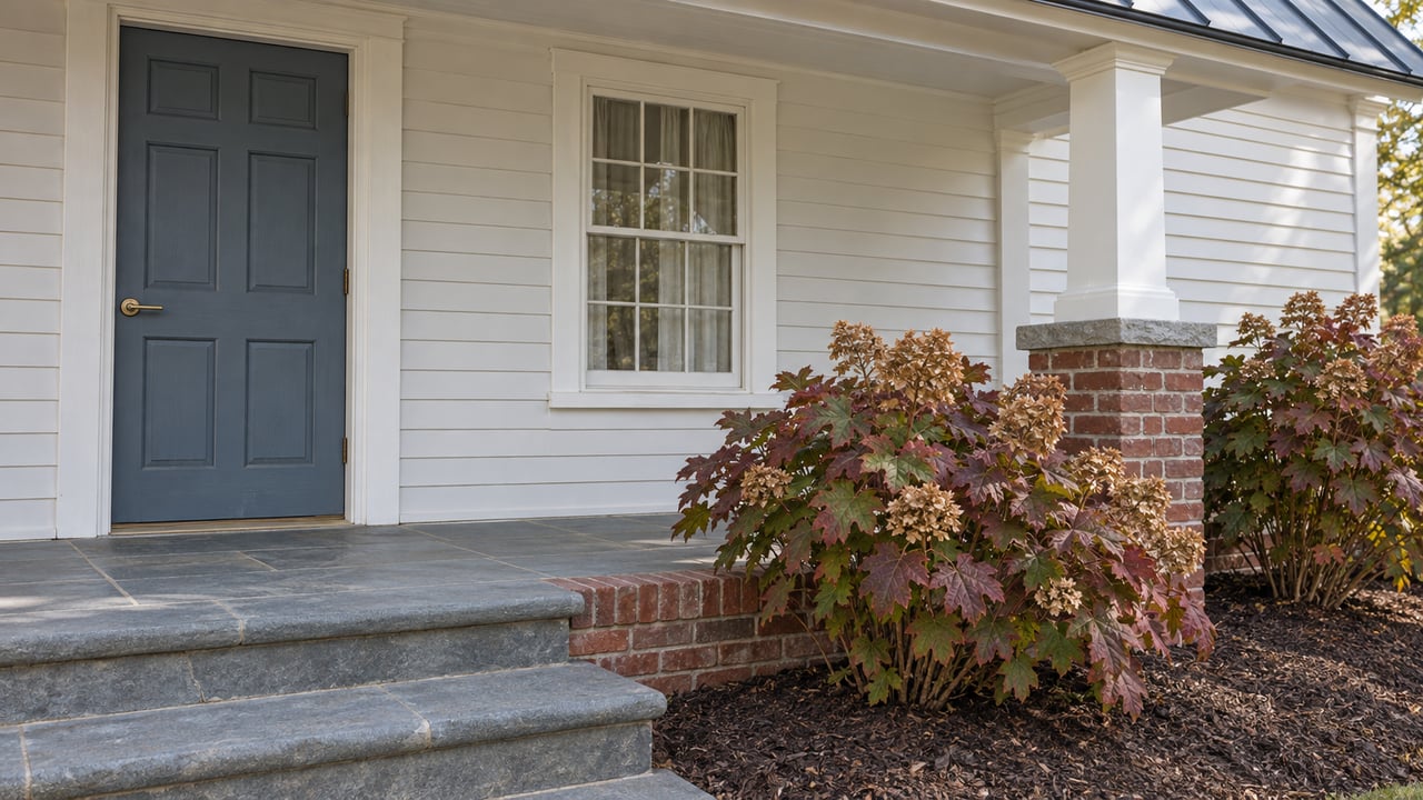Clapboard home entrance with a slate-blue door, bluestone steps, brick foundation and autumn oakleaf hydrangeas.
