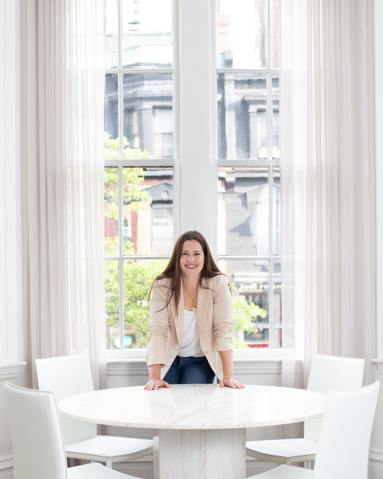 Claire McClintock in a cream blazer leans on a round table in front of tall windows.
