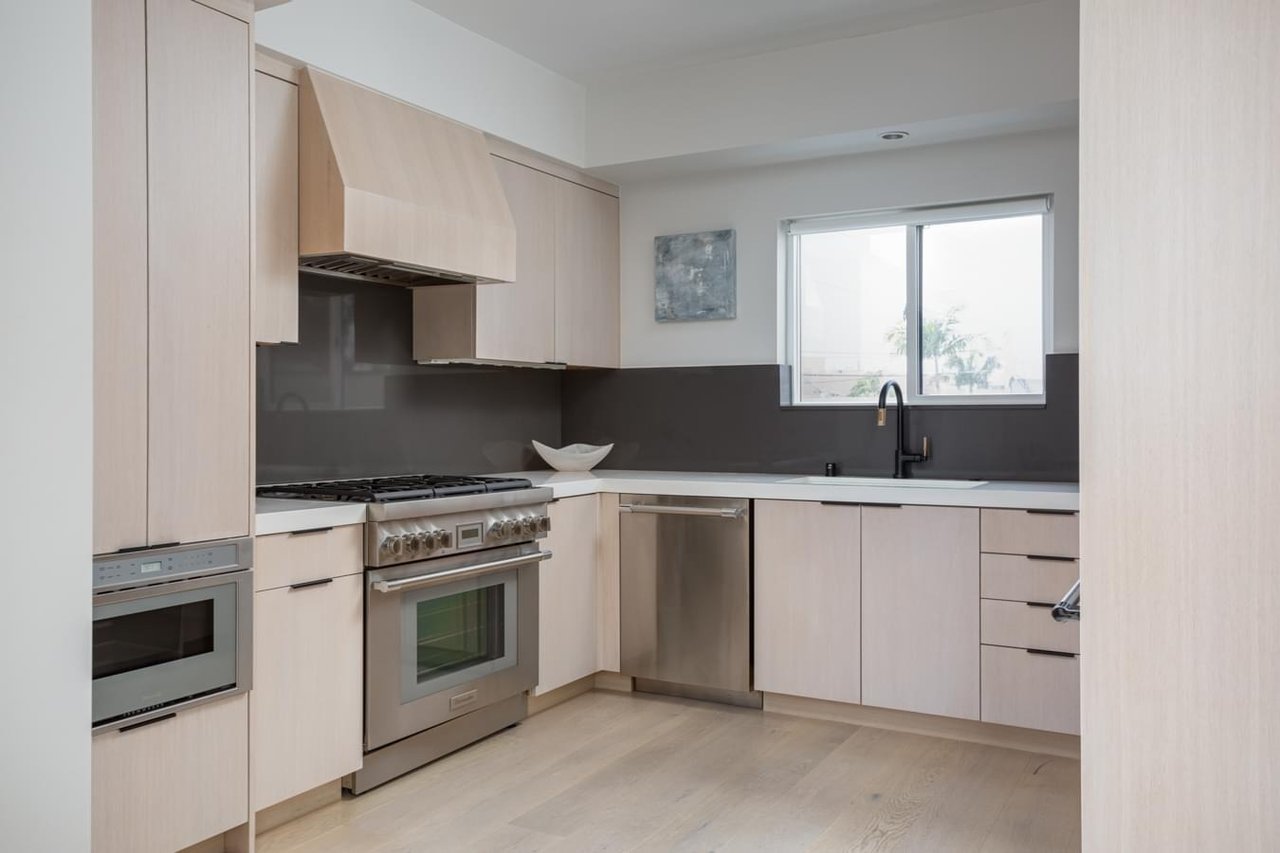  A contemporary kitchen with light wood cabinets, stainless steel appliances, and a gray backsplash with a window above the sink.