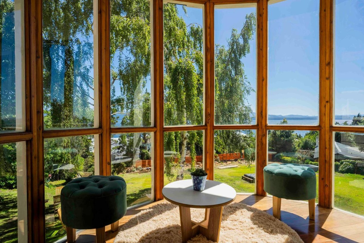 Sunroom with tall wood-framed windows overlooking a garden and lake, furnished with green chairs and a round table.