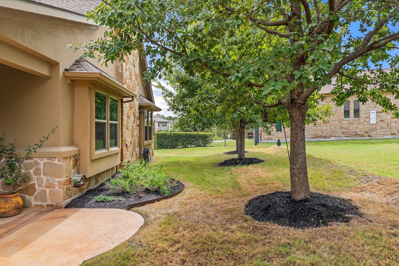 Side yard with mature shade trees beside a stone-and-stucco house and curved patio.