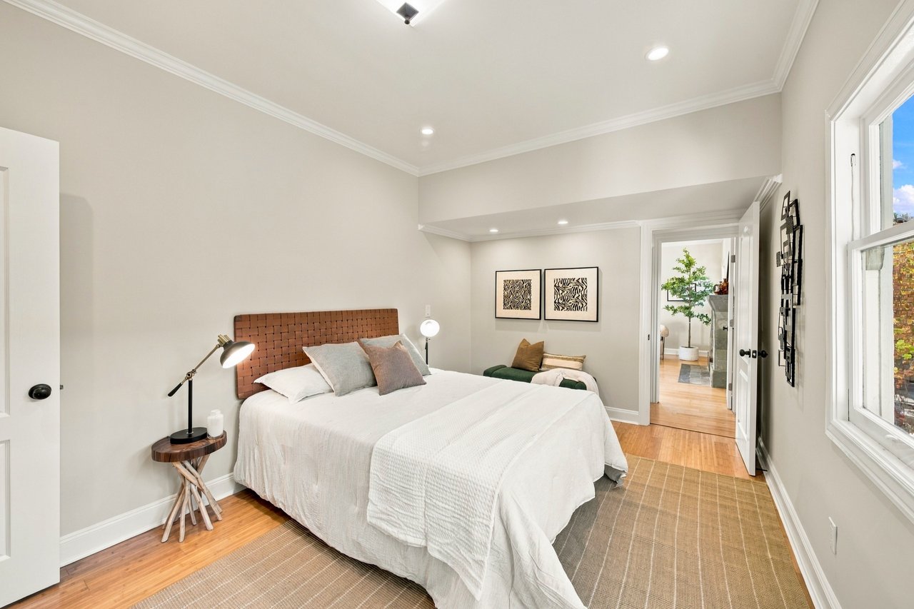 Staged bedroom with a large white bed, woven headboard, hardwood floors, and a wide window.