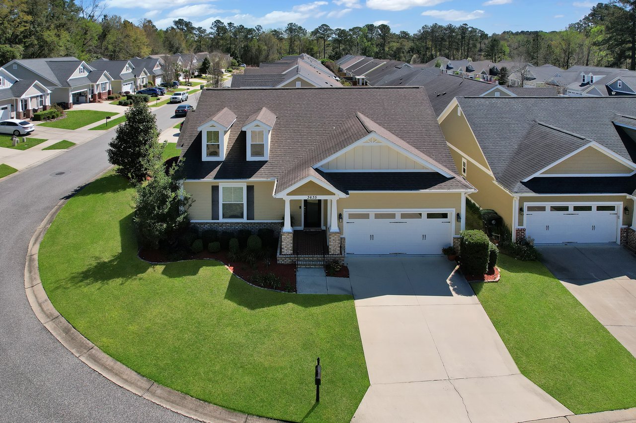 Aerial view of a suburban neighborhood with neatly aligned houses. A prominent beige house with a dark roof, manicured lawn, and double garage is centered.