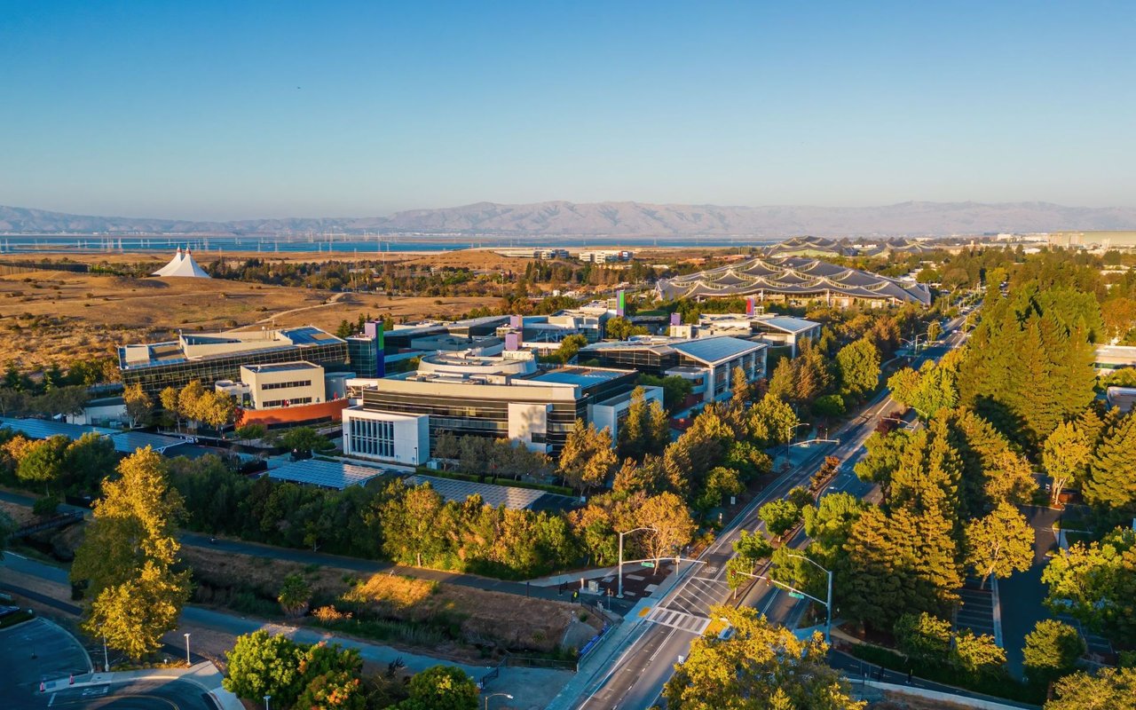 Aerial view of Mountain View office buildings, tree-lined streets, bay, and distant mountains.