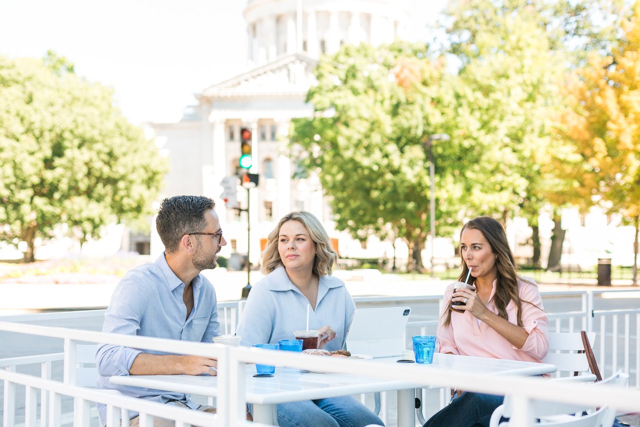Cait Berry of Insiders Realty meeting with two homebuyers at an outdoor cafe table with the Wisconsin State Capitol visible in the background, discussing their Madison home search over coffee.