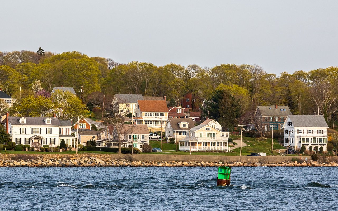 Waterfront neighborhood of houses along a rocky shore, with a green navigation buoy in the foreground.