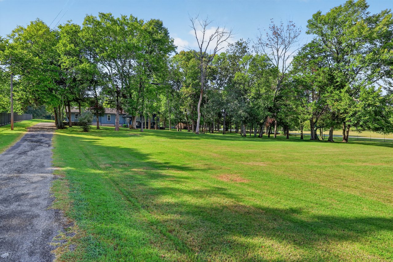 Tree-lined lawn and gravel driveway leading to a partially obscured blue house.