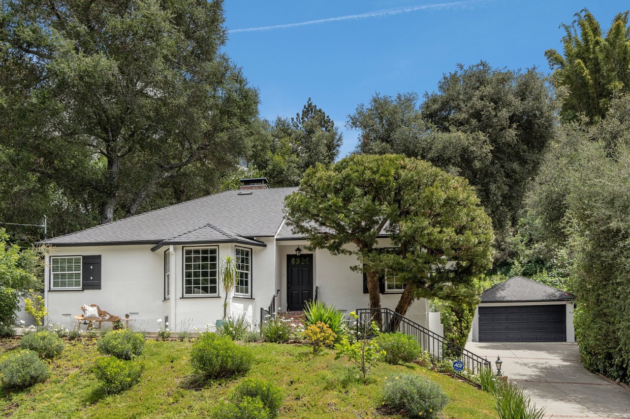 White single-story house with a bay window, black front door, mature trees, and a garage beside the driveway.