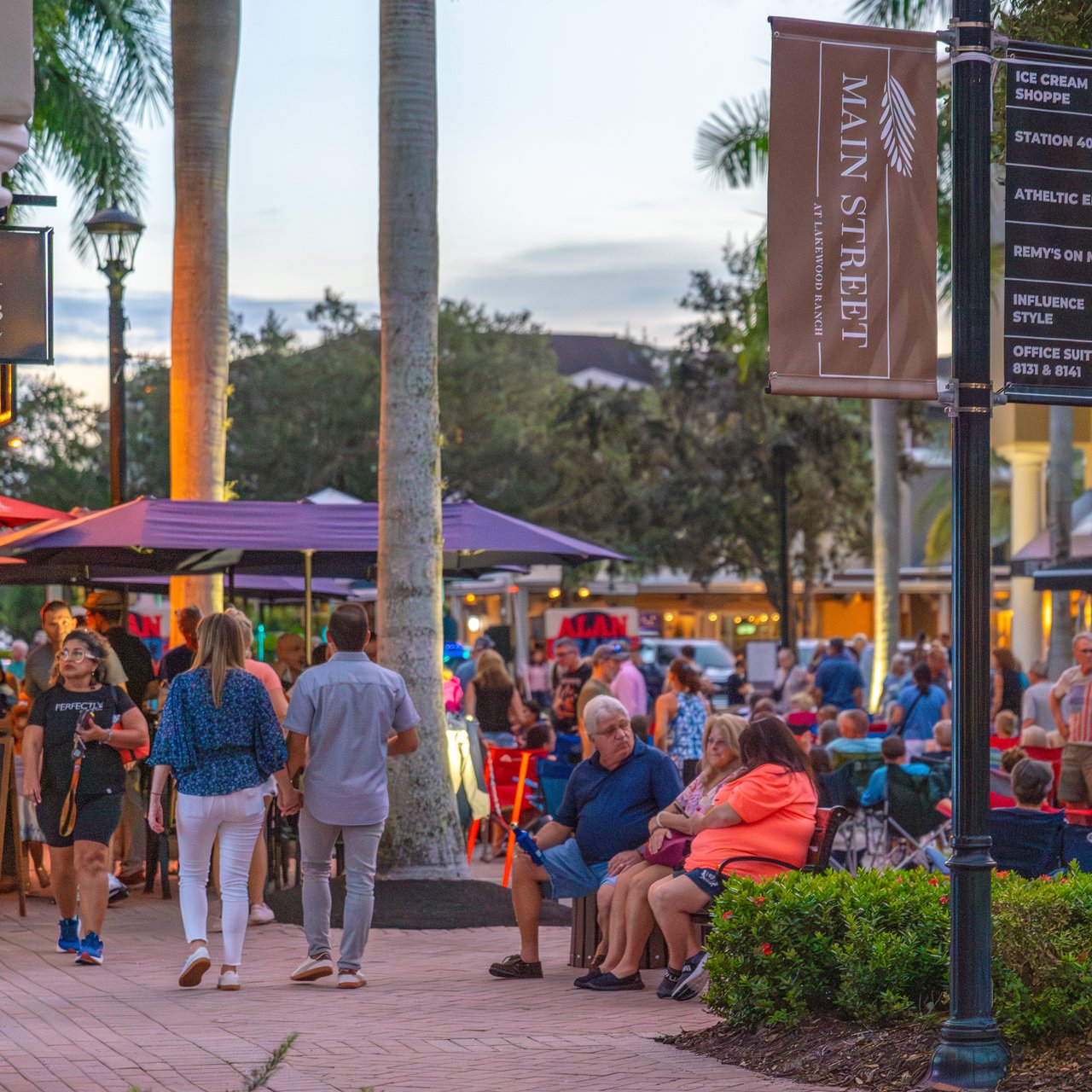 Lakewood Ranch Main Street at dusk, with palm-lined shops, outdoor dining, and a busy pedestrian walkway.