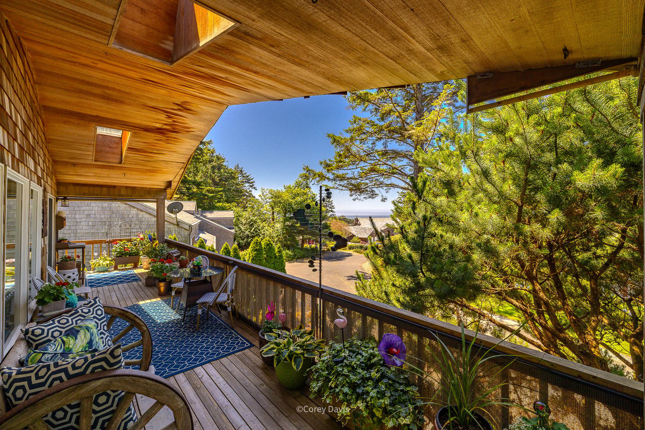 Covered deck in Cannon Beach, Oregon, with outdoor seating, potted plants, trees, and a partial ocean view in the distance.