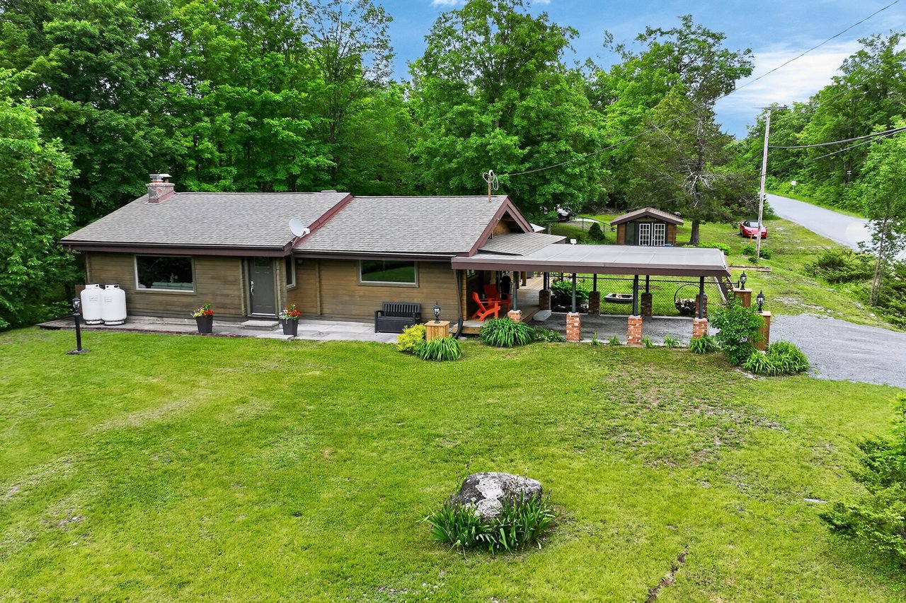 Single-story wood-sided home with a covered carport, orange patio chairs, and a broad green lawn.
