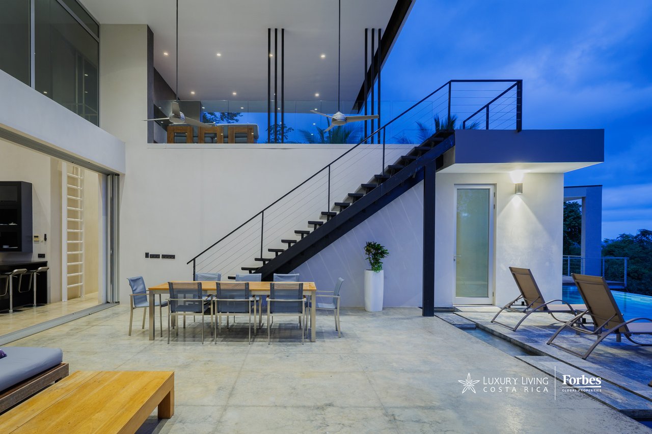 Modern home patio with outdoor dining, a striking staircase, and lounge chairs beside the pool at dusk.
