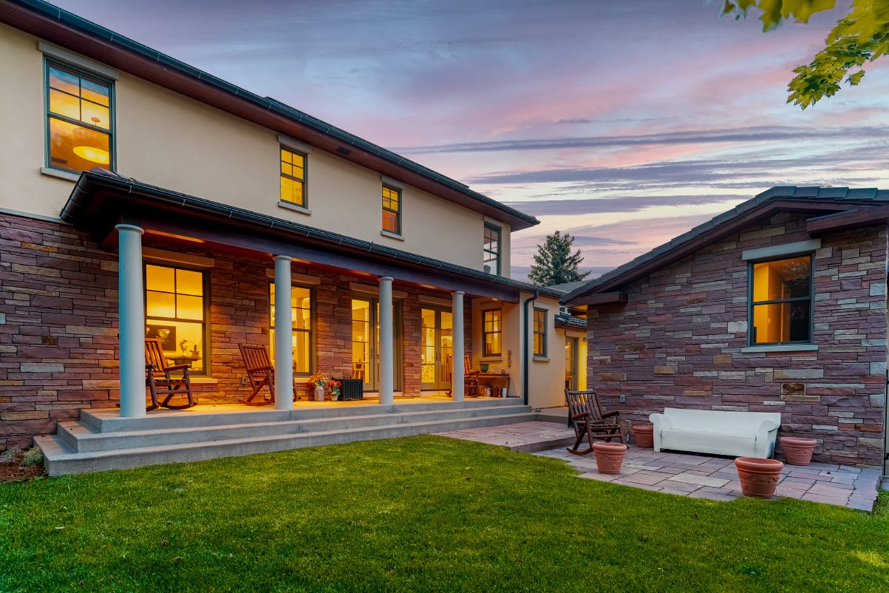 Back porch view at dusk with glowing windows, stone facade, white columns, and pink sunset sky