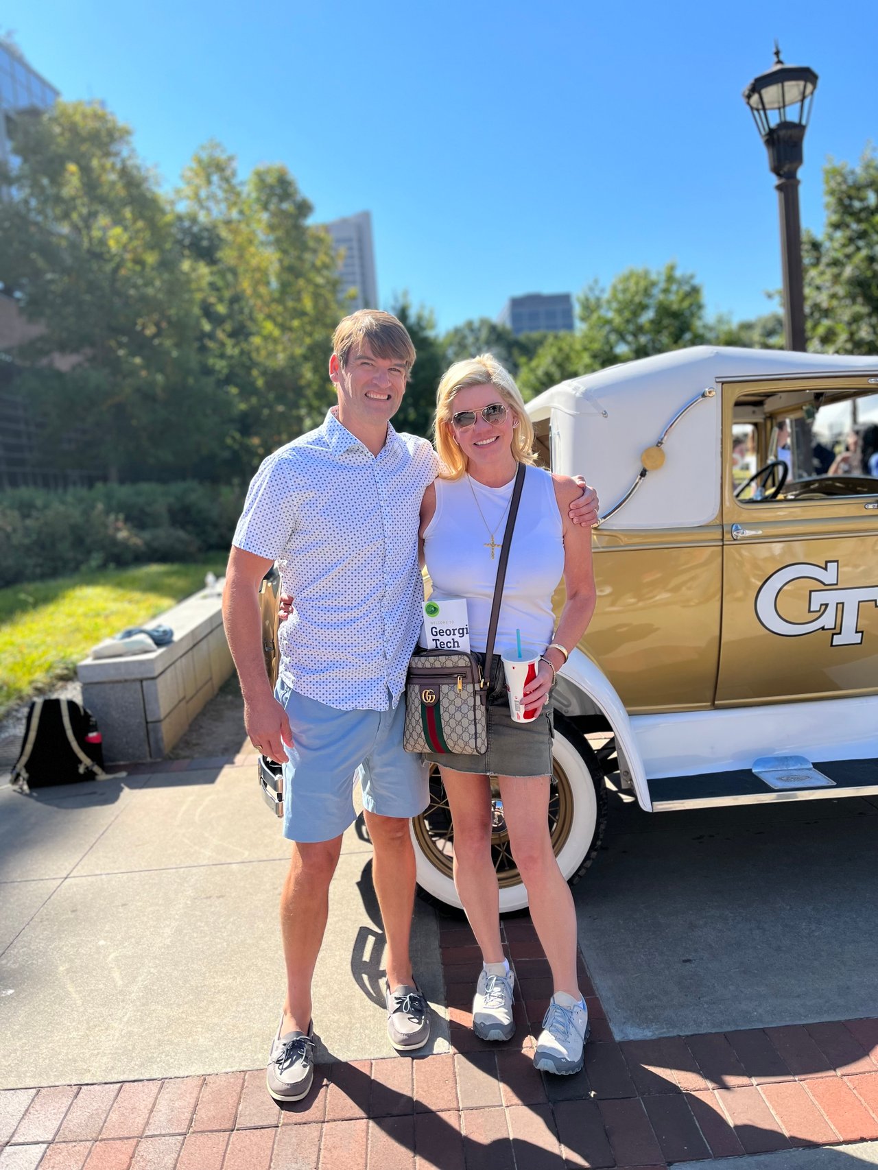 A man and woman pose beside a gold-and-white vintage car on a sunny sidewalk.