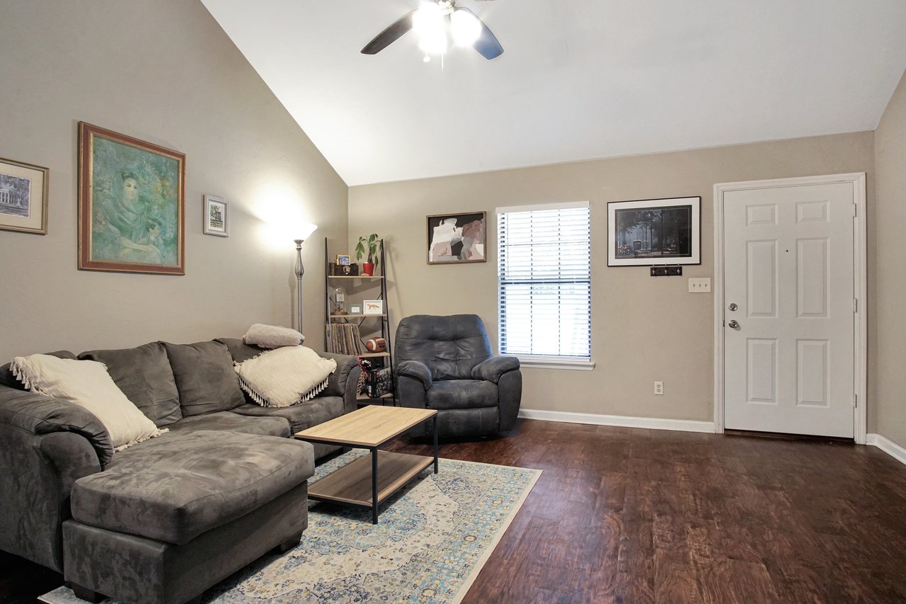 Cozy living room with gray sectional sofa, reclining chair, and wooden coffee table on a patterned rug. Walls feature framed art, lit warmly by a floor lamp.