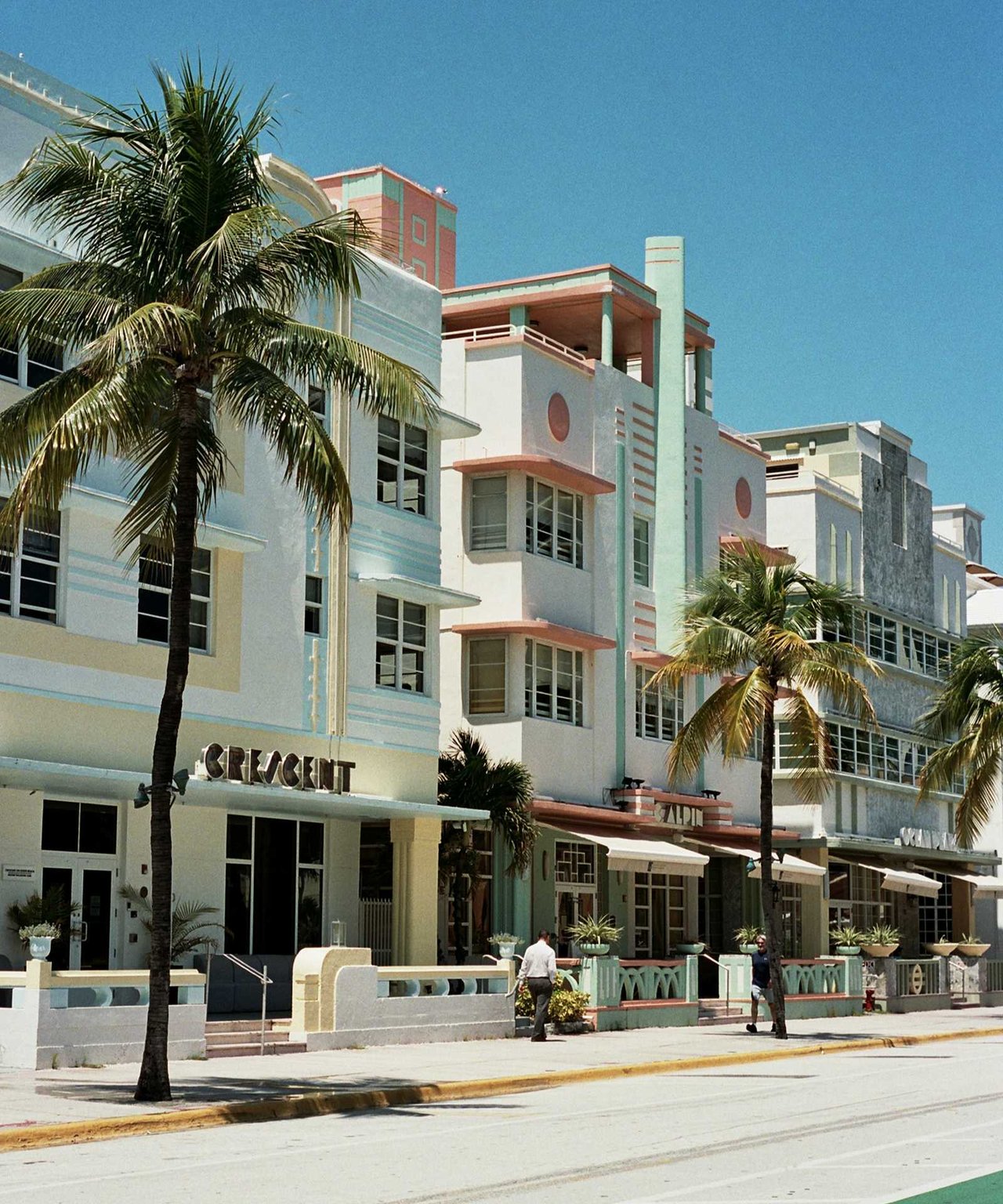 Pastel Art Deco storefronts with geometric facades and palm trees along a sunny sidewalk.