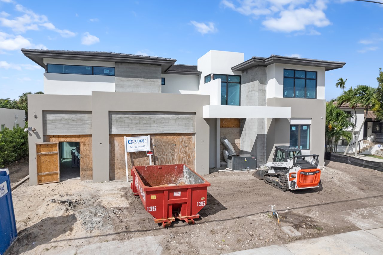 Modern two-story home under construction with plywood-covered garage doors and construction equipment in the driveway.