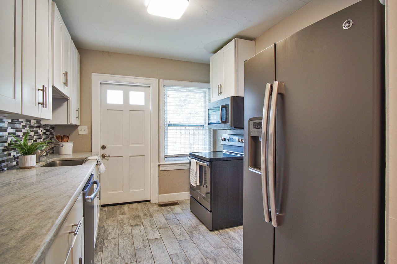 Compact kitchen with stainless steel fridge, oven, and microwave. White cabinets, a potted plant, and a colorful tile backsplash. Bright and inviting.