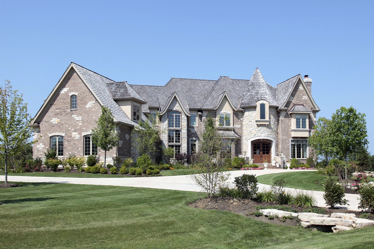 Large stone-and-brick house with a conical tower, arched entrance, and landscaped front lawn.
