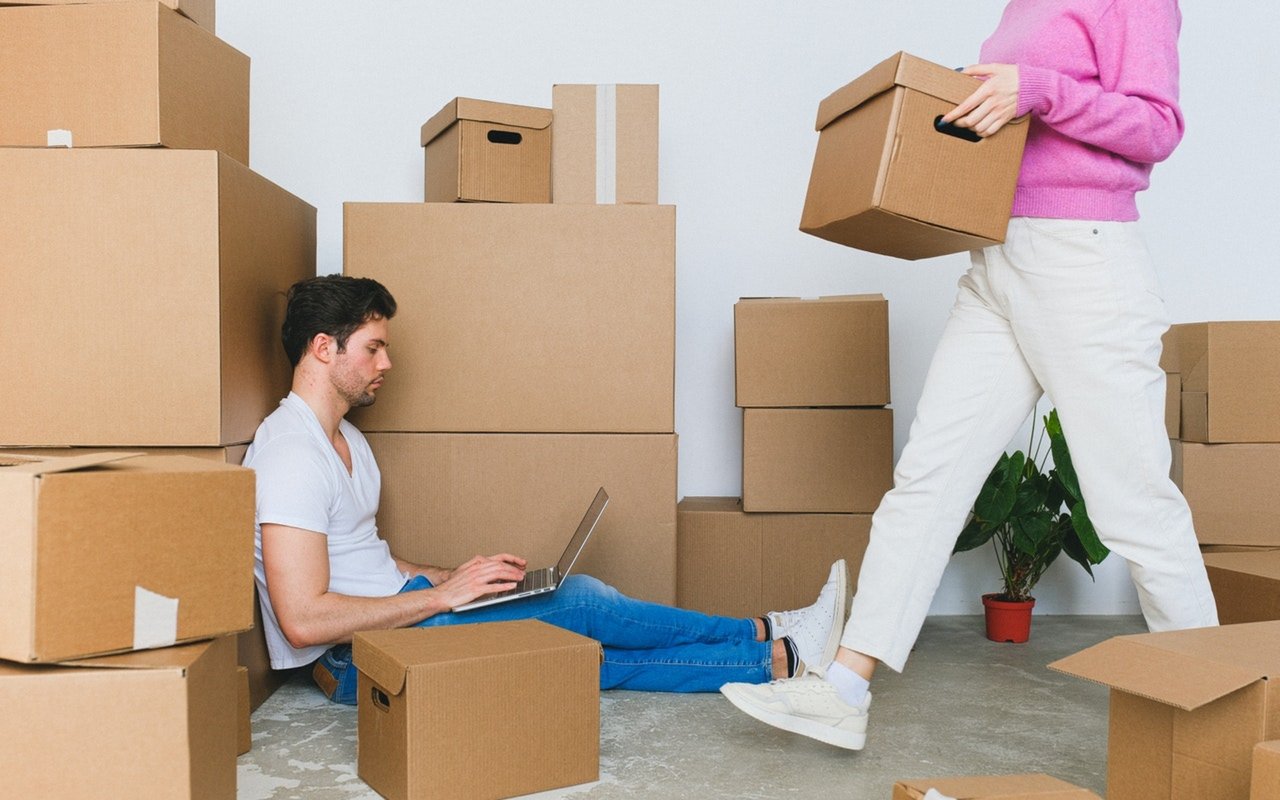 A couple move among cardboard boxes as the man works on a laptop and the woman carries a box.