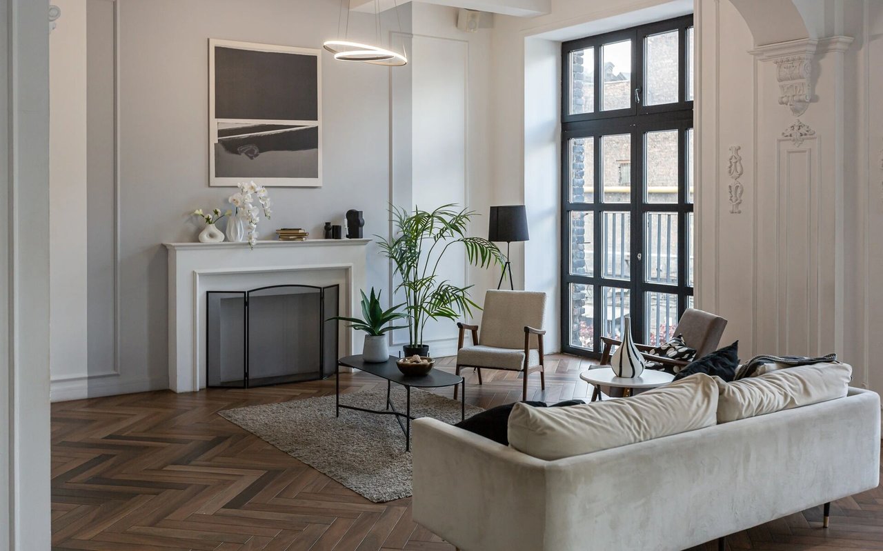 Living room with a fireplace, herringbone wood floors, and tall black-framed windows.