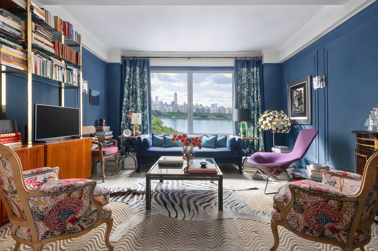 Vibrant blue maximalist living room featuring floor-to-ceiling bookshelves, a zebra-print rug, and patterned armchairs, centered around a large window framing a scenic view of the reservoir and city skyline.