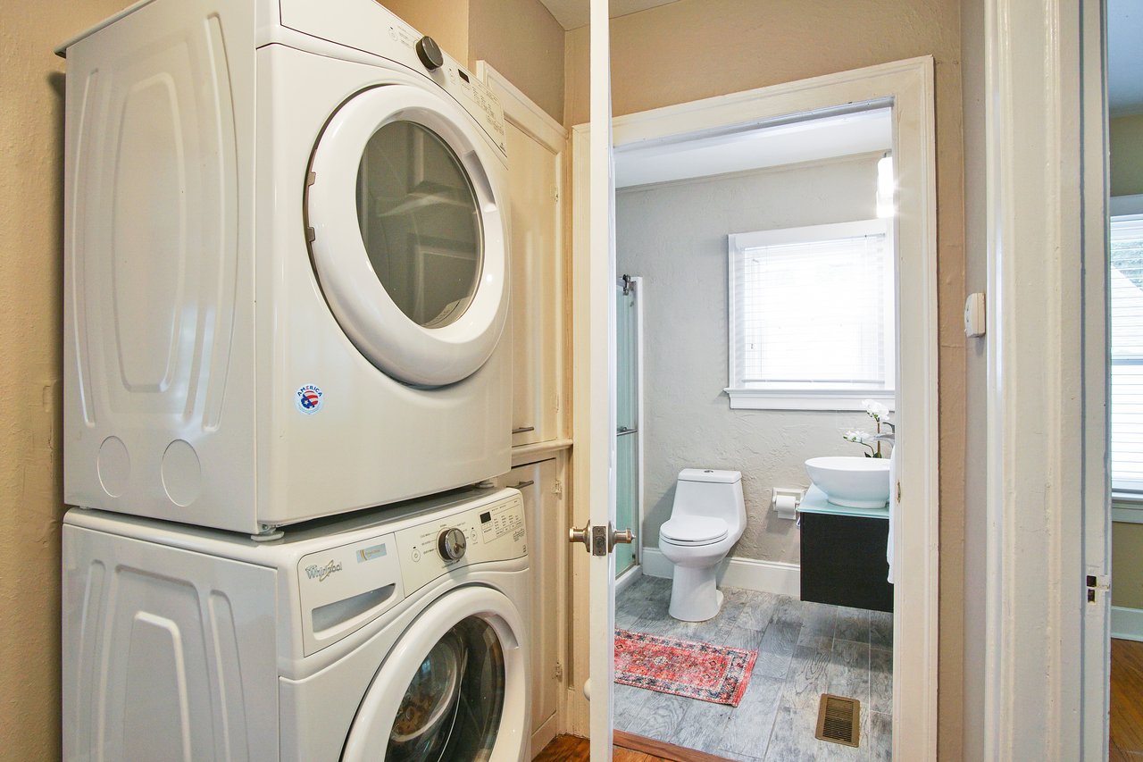 Stacked washer and dryer in a narrow closet, leading to a small bathroom with a white toilet, vessel sink, and a window. Bright, clean space.