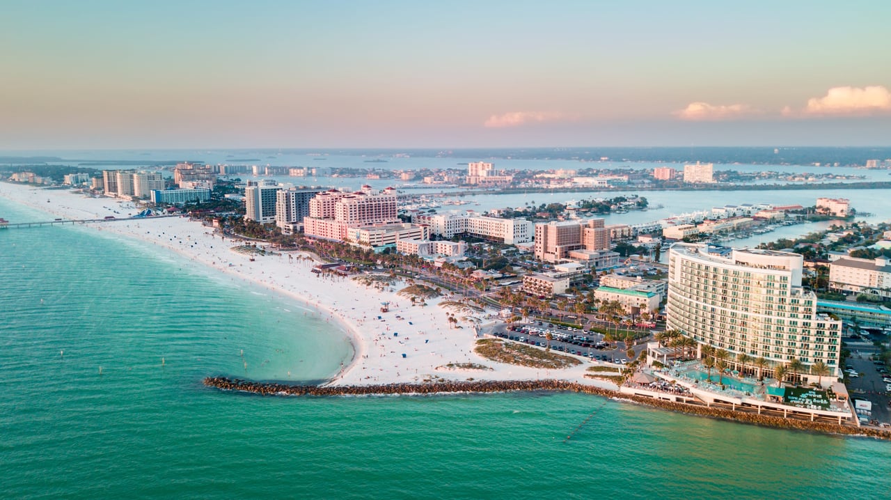  An aerial view of Clearwater Beach, Florida, showcasing a long stretch of white sandy beach packed with people and colorful beach umbrellas.