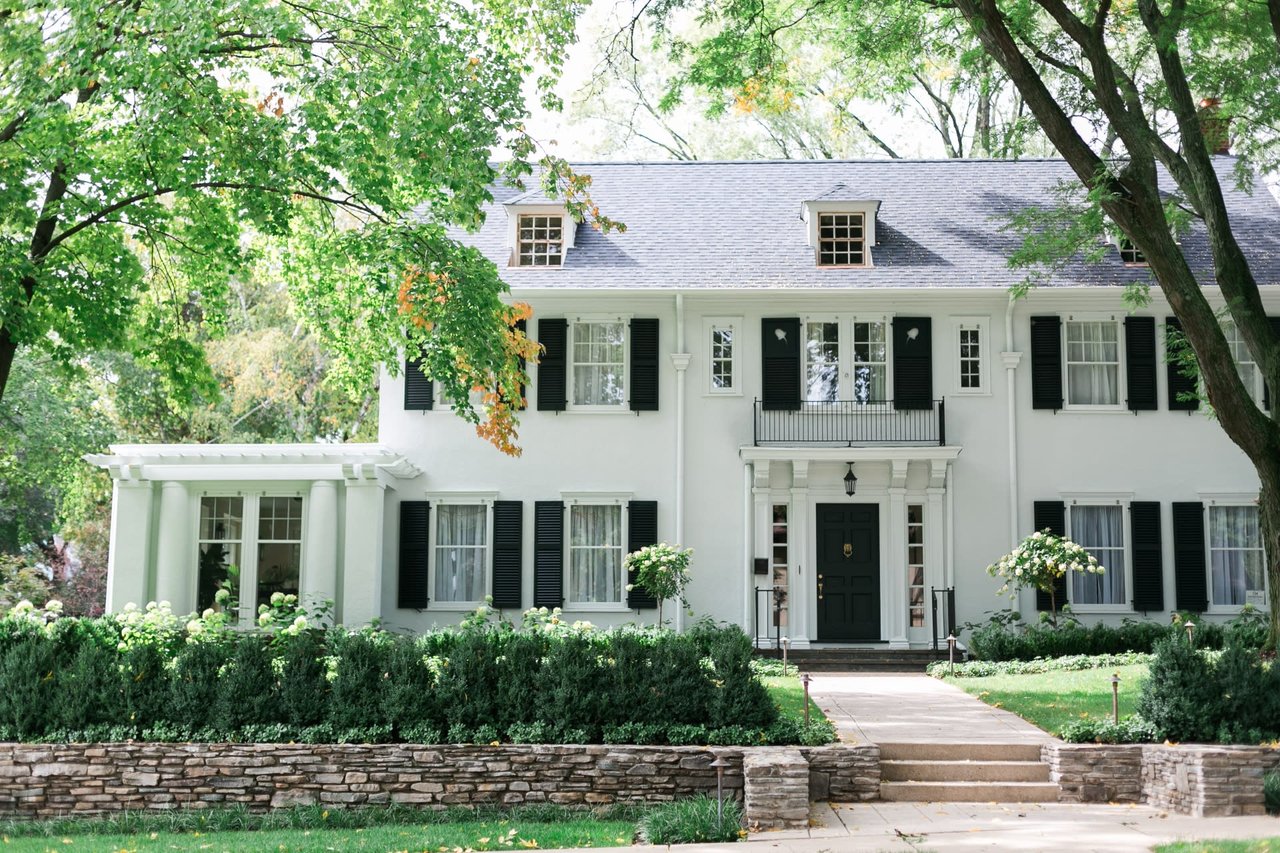 A white house with black shutters, a stone planter with bushes, a walkway to the front door, a lawn, and surrounded by trees.