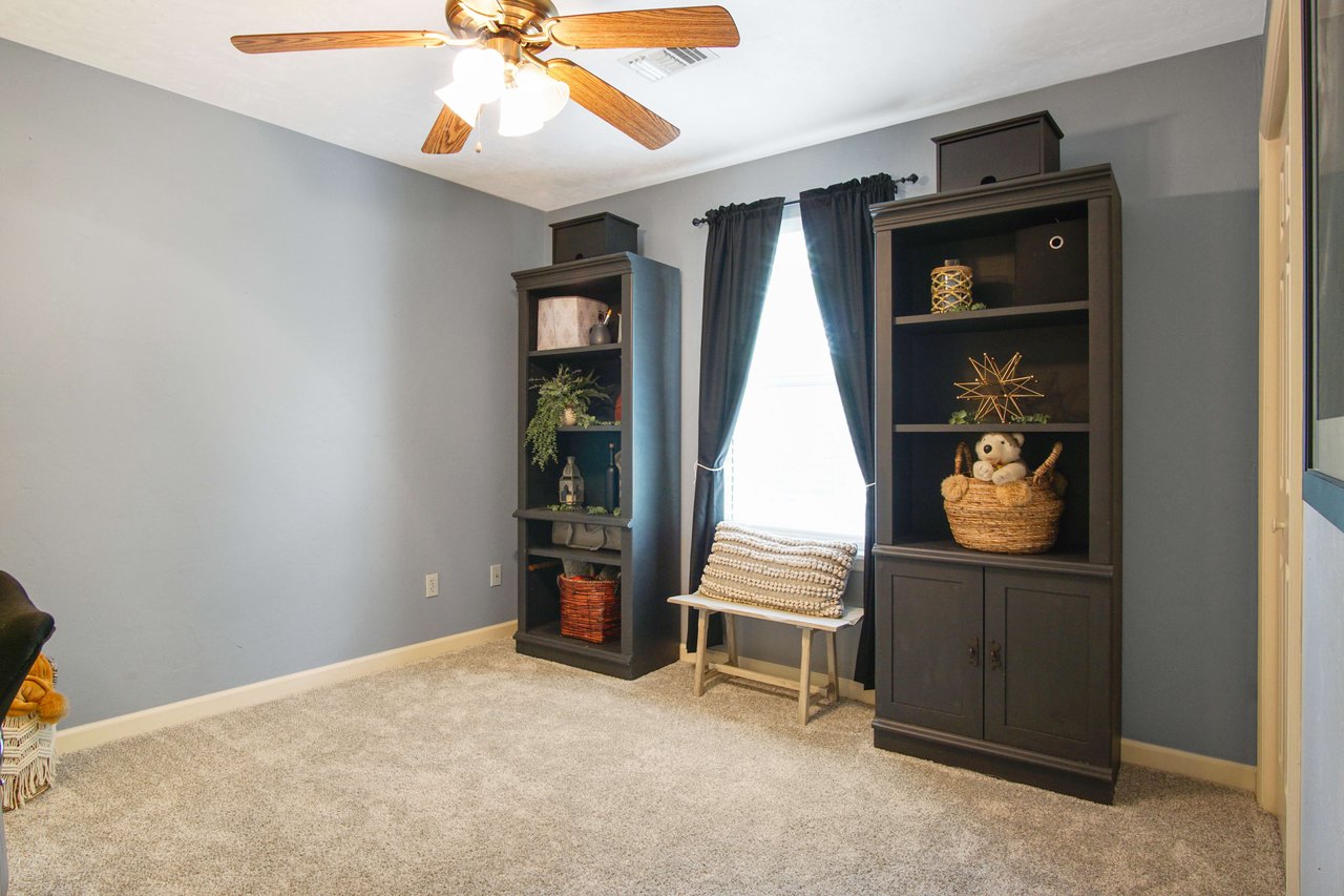 An interior shot of a bedroom with blue walls, carpeted flooring, and a ceiling fan. The room includes a dresser and a window with curtains, creating a bright and inviting space.