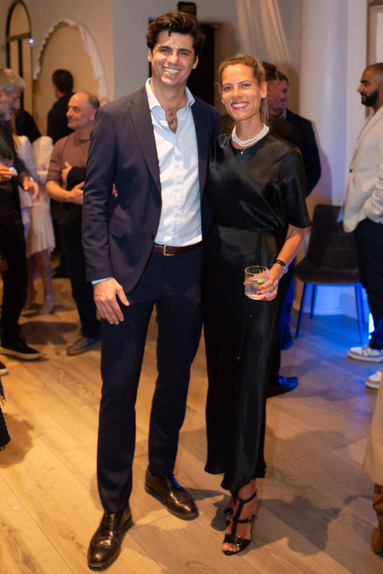 A man in a navy suit and a woman in a black dress pose together at a gathering.