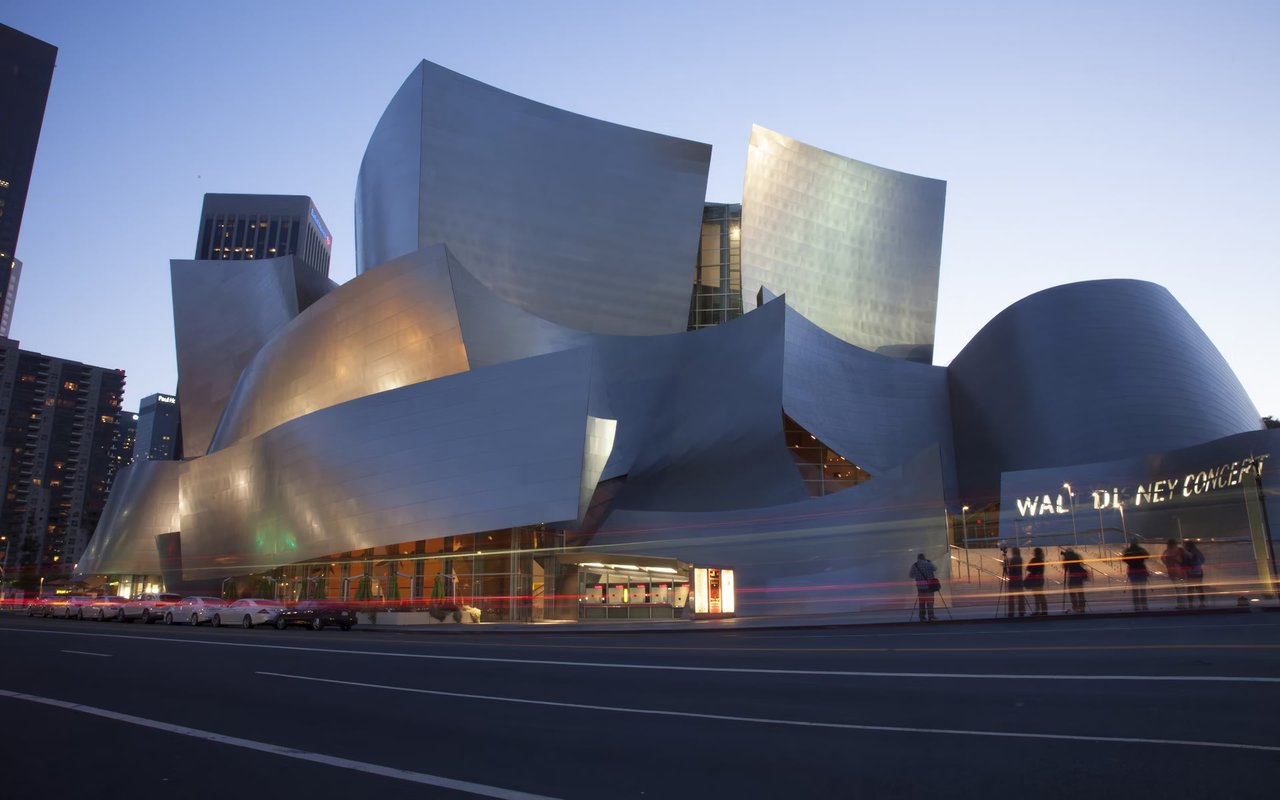 Walt Disney Concert Hall in Los Angeles, with its unique design, illuminated at night, with people and parked cars.