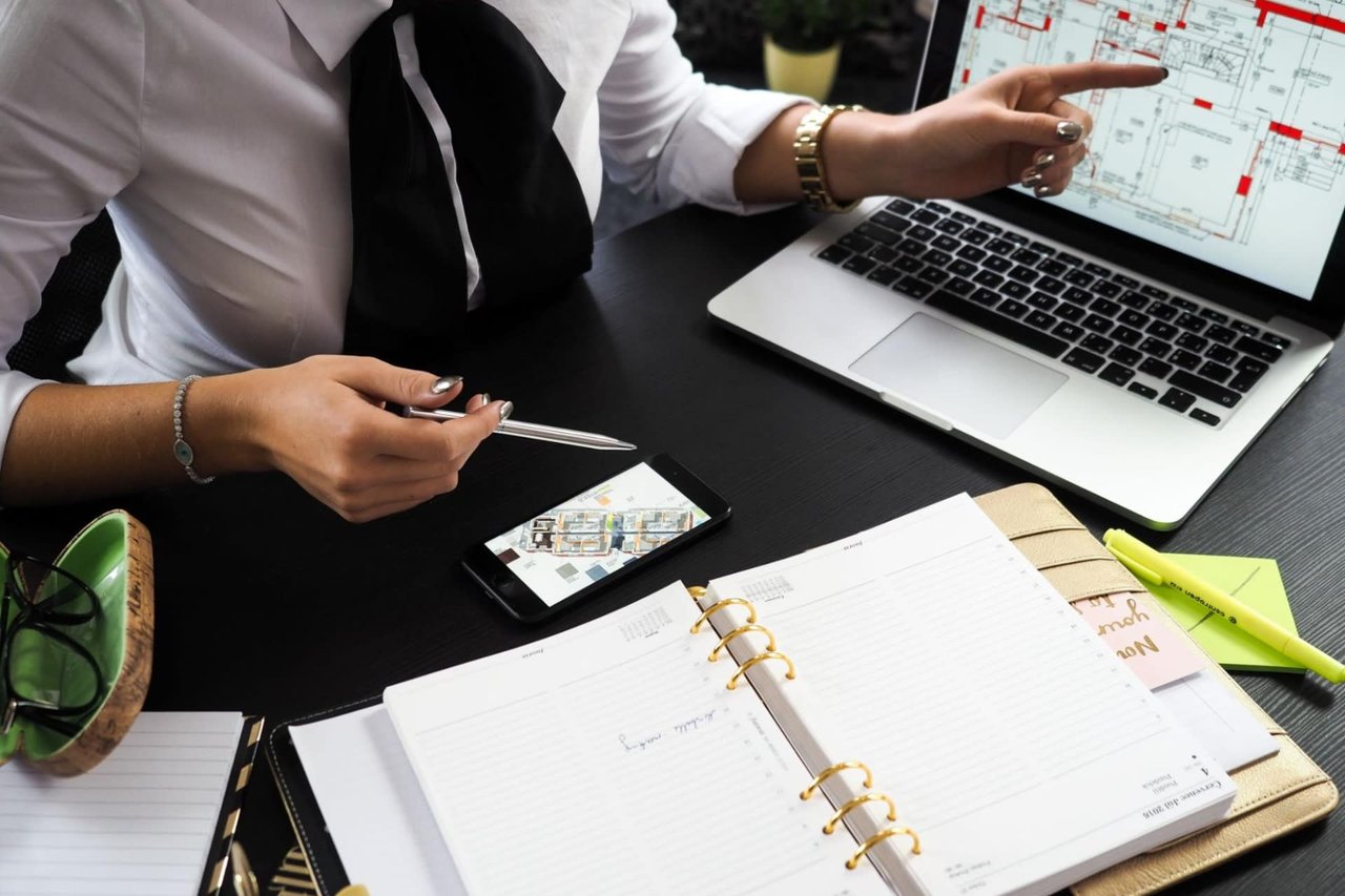  A person wearing a white shirt with a black tie and a gold watch is seated at a dark desk, pointing at a laptop screen that displays architectural plans or blueprints.