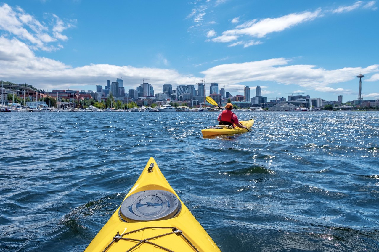 A person on a yellow kayak floating on a calm lake, with a city skyline in the background.