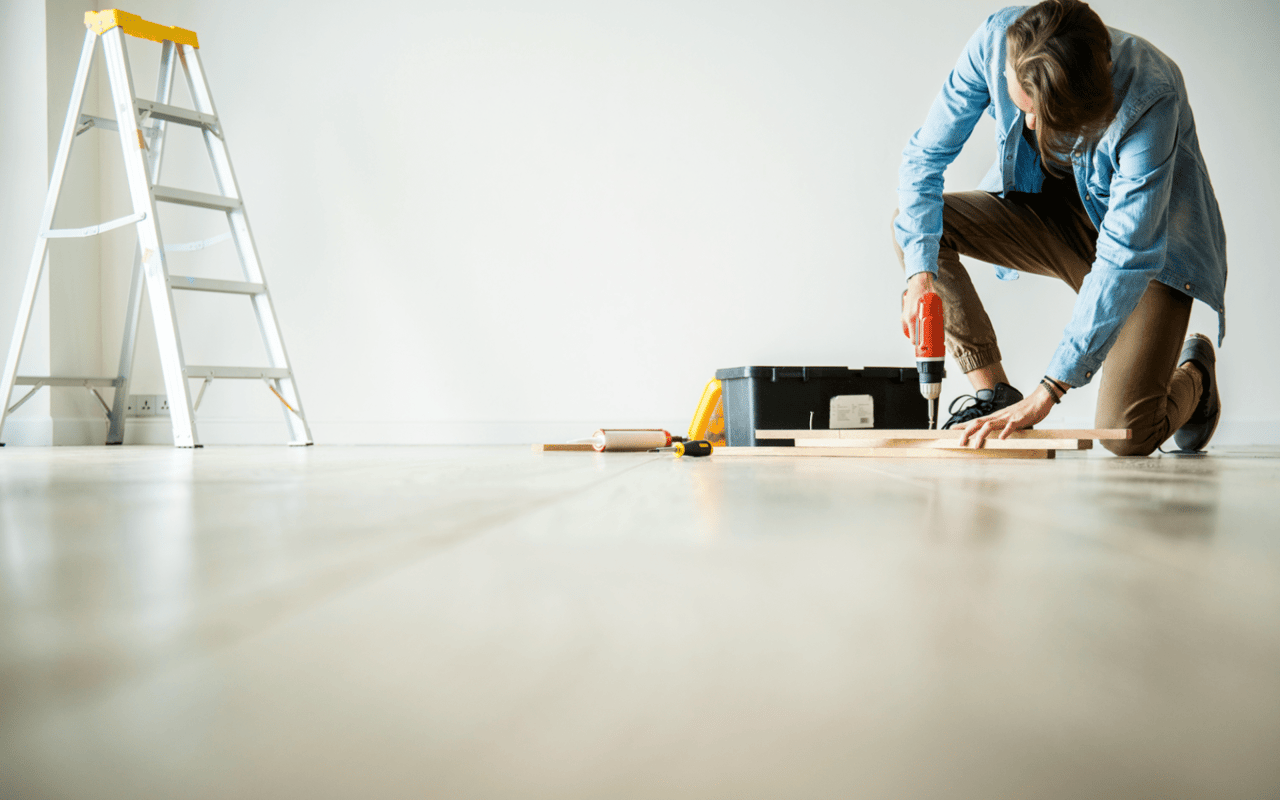  A person wearing a blue long-sleeved shirt and brown pants is kneeling on a light wooden floor, focused on drilling into a piece of wood
