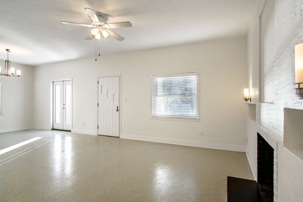 An interior shot of an empty living room with white walls, tiled flooring, and large windows. The room appears clean and spacious with modern light fixtures.