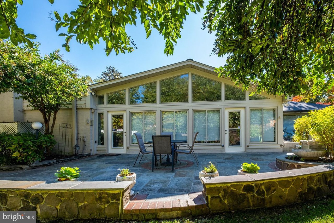 Home’s rear glass wall and stone patio with outdoor dining furniture, framed by mature trees.