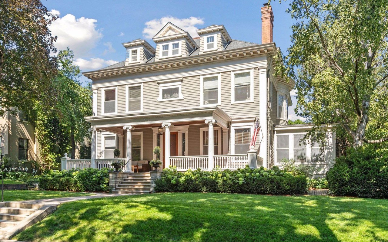 A large, two-story house with a wraparound porch and a white picket fence with well-kept lawns and trees.