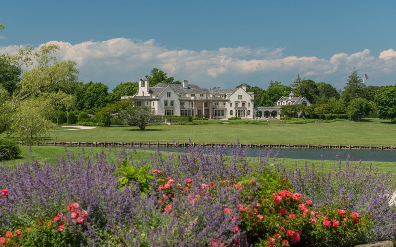 A large white house on a green field with lavender, catnip, a pond with a deck, and distant surrounding trees.