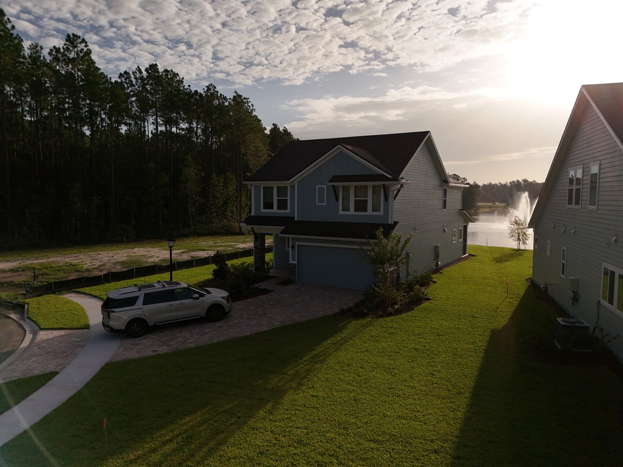 Two-story blue-gray home beside a lake, with a driveway, white SUV, and fountain.