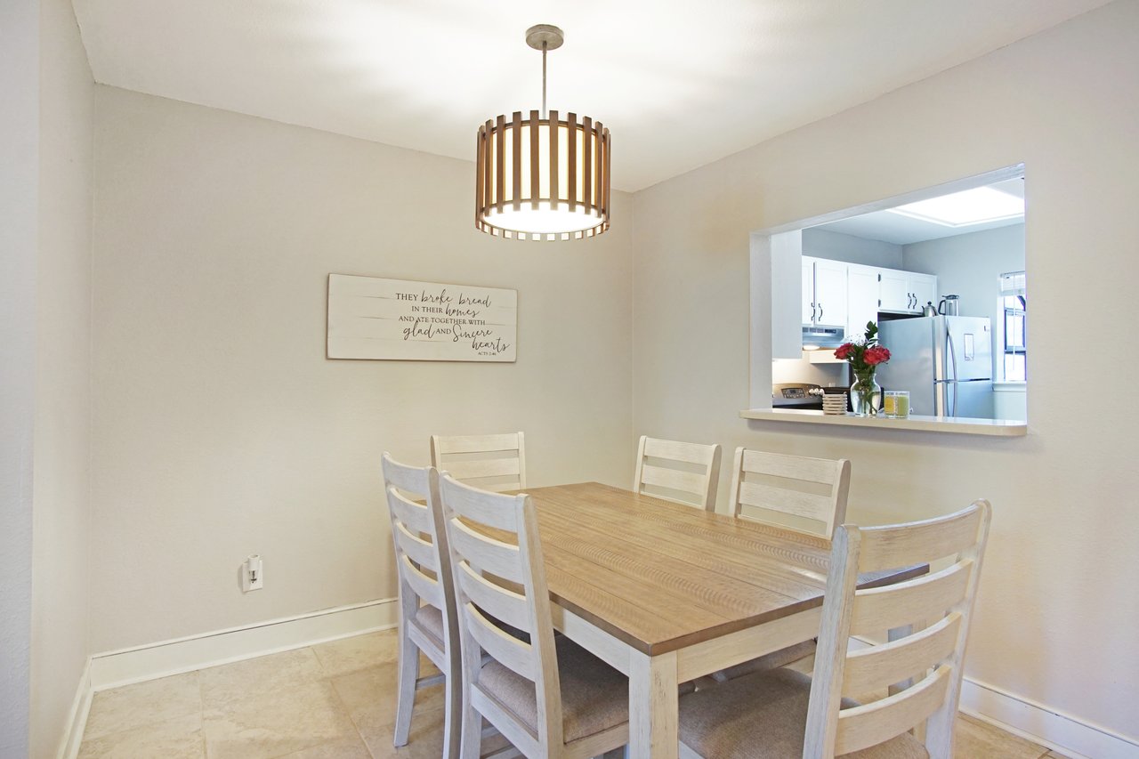 Cozy dining area with a wooden table and six chairs under a modern pendant light. A sign on the wall and a pass-through to a bright kitchen add a welcoming touch.