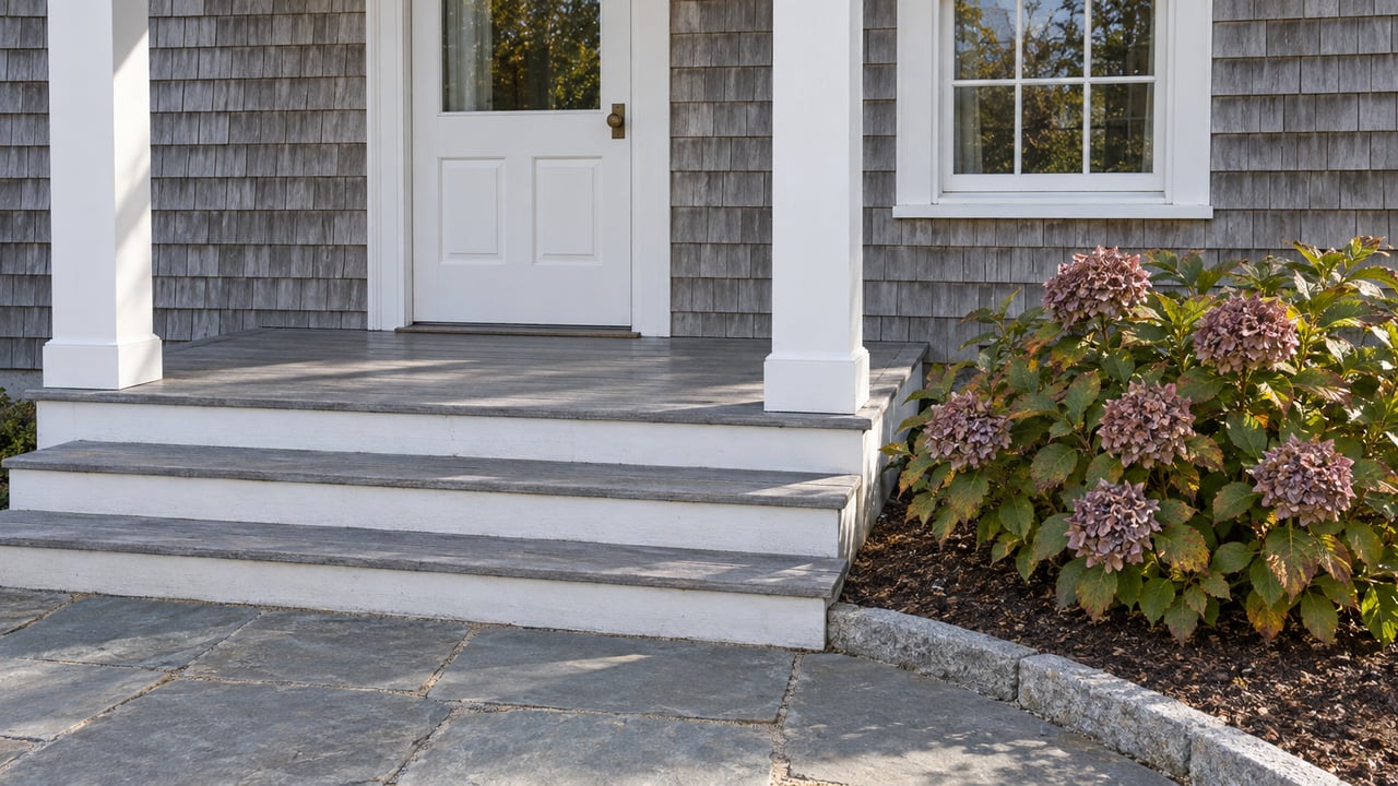 Weathered cedar-shingle entry with white trim, gray porch steps, bluestone paving and faded hydrangea blooms in autumn daylight.