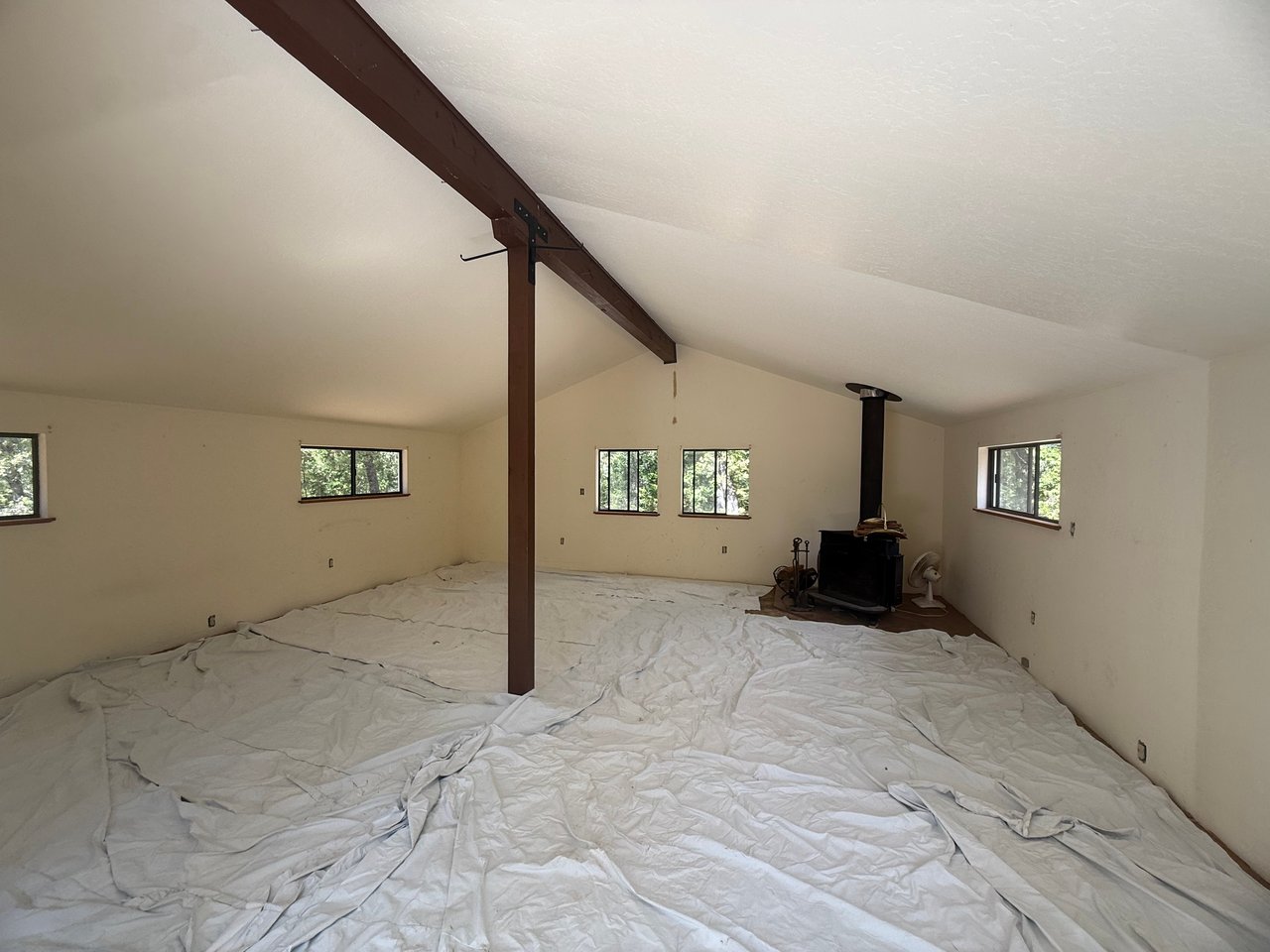 Vacant vaulted room with exposed beams, multiple windows, and a wood stove; protective sheeting covers the floor.