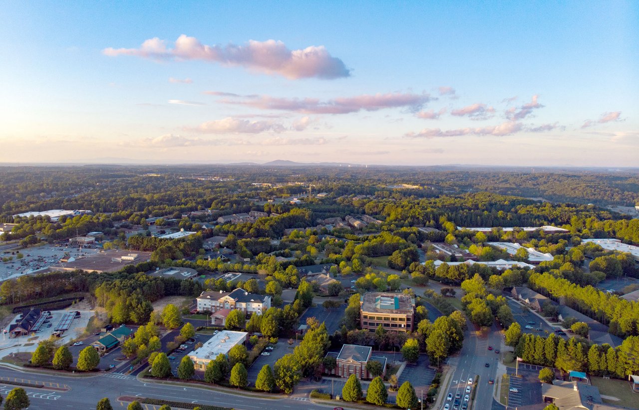 Aerial view over tree-lined neighborhoods and commercial buildings beneath a pastel evening sky.